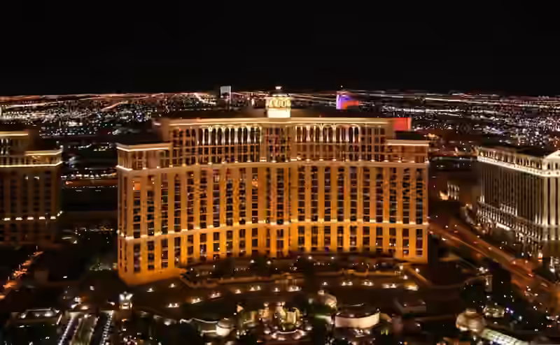 The curved main building of Bellagio lit at night, seen from above, with the low roofs of the resort's ground level in front and the lights of Las Vegas behind