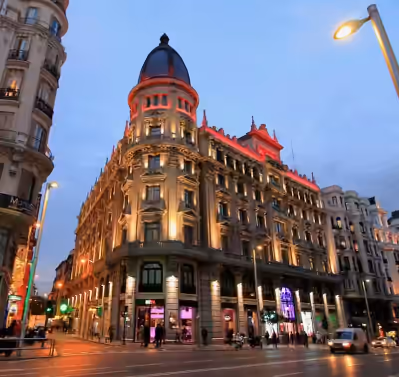 The corner building at Gran Vía 24 in Madrid at dusk, lit from below, with a round tower topped by a dark slate dome