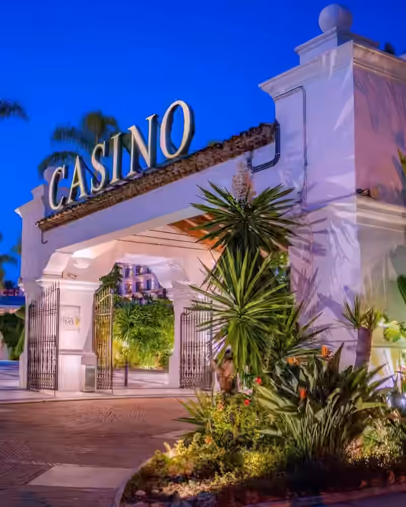White gateway with a tiled roof and large letters spelling Casino, the entrance to Casino Marbella at dusk, with palms and garden plants in front