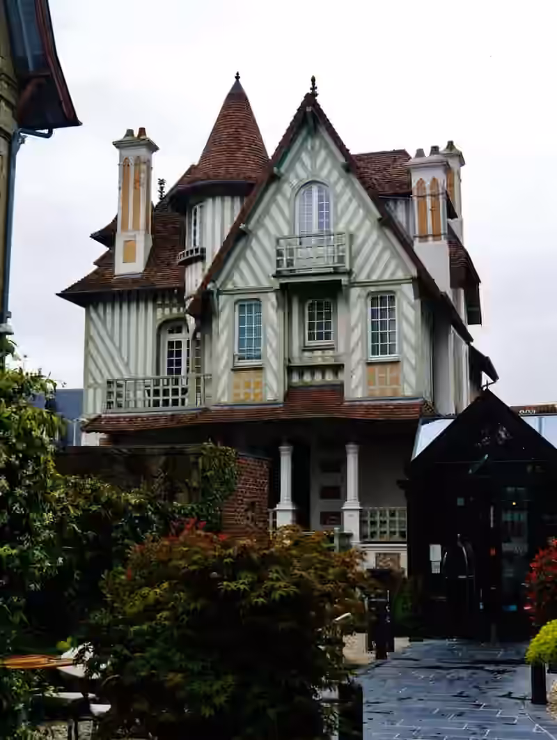A turreted half-timbered villa of the Villa Augeval hotel, seen from the garden path beside a glazed entrance