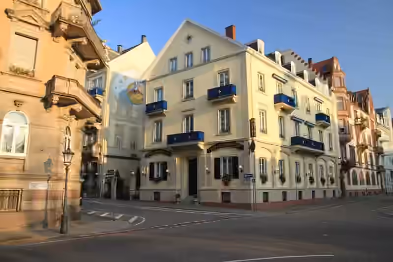 Hotel Der Kleine Prinz in Baden-Baden, a pale yellow corner building with blue balconies, next to a wall painting of the Little Prince