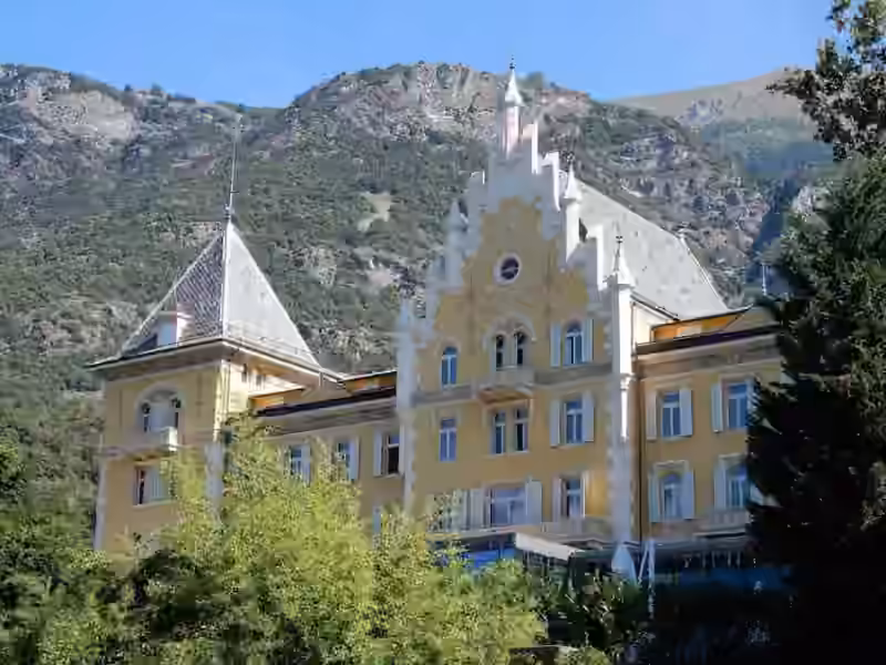 The yellow facade of Grand Hôtel Billia in Saint-Vincent, with a white stepped gable and a slate-roofed tower, seen above trees with a mountainside behind