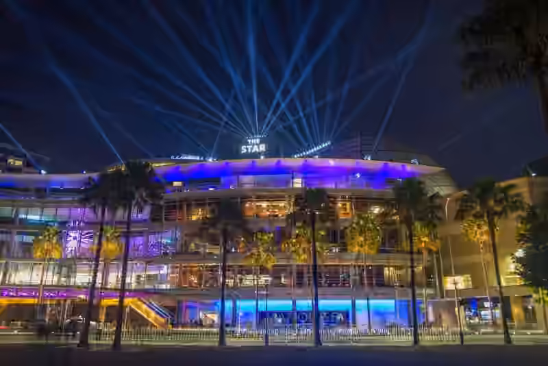 The glass front of The Star Sydney at night, lit in blue and purple behind a row of palm trees, with light beams above the sign on the roof