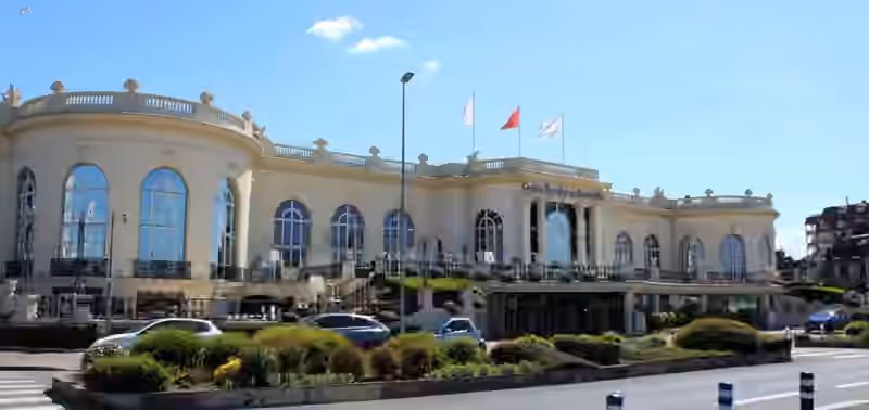 The cream-coloured main facade of Casino Barrière de Deauville, with its name above the entrance, tall arched windows, a terrace and three flags on the roof